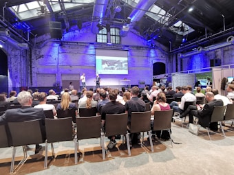 A conference room filled with attendees seated and facing a stage where two speakers stand in front of a presentation screen displaying an image. The room has high ceilings with industrial architecture and blue lighting.
