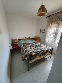 A small bedroom with a wrought iron bed featuring a floral-patterned bedspread occupies the center. The walls are painted white, and a simple brown lampshade hangs from the ceiling. Two wooden side tables flank the bed, with personal items placed on them. Lace curtains cover a window on the right, allowing some natural light to enter. A few photos or small artworks are hung on the wall near the bed.