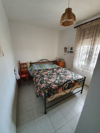 A small bedroom with a wrought iron bed featuring a floral-patterned bedspread occupies the center. The walls are painted white, and a simple brown lampshade hangs from the ceiling. Two wooden side tables flank the bed, with personal items placed on them. Lace curtains cover a window on the right, allowing some natural light to enter. A few photos or small artworks are hung on the wall near the bed.