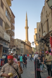 A vibrant street scene features a tall minaret rising in the background, surrounded by historic buildings and bustling activity. People in varied attire are walking, shopping, and engaging with street vendors. The cobblestone street adds to the traditional atmosphere, and shops with Arabic signage line the sides.