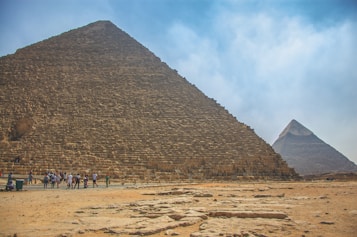 Large ancient pyramids dominate the landscape, with a group of tourists walking along the sandy base. The sky is clear with a few clouds, creating a majestic and historical atmosphere.