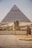 A large, ancient pyramid stands prominently in the background, with the iconic Great Sphinx of Giza situated in front of it. The stone structures are surrounded by a sandy landscape and a paved area, with some visitors and a person near a fence observing the historical site.