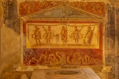 A detailed fresco with classical figures holding objects and raising hands, depicted in a rectangular panel framed by ornate architectural elements. The figures are set against a faded background, with elaborate decorative motifs including serpentine shapes below.