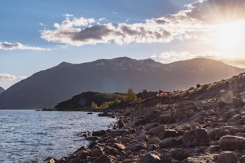 A serene lakeside scene with rocky shores and gentle waves. The background features majestic mountains capped with patches of snow. The sky is partly cloudy with the sun low on the horizon, casting warm light and lens flares. There are tents set up along the grassy area near the water, indicating a campsite.