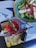 A vibrant green salad served in a blue bowl contains lettuce, sliced onions, feta cheese, and a caper. Next to the bowl, there's a red drink with a sprig of rosemary on top. A small, dark dropper bottle is positioned nearby, all placed on top of an open magazine with colorful pages.