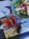 A vibrant green salad served in a blue bowl contains lettuce, sliced onions, feta cheese, and a caper. Next to the bowl, there's a red drink with a sprig of rosemary on top. A small, dark dropper bottle is positioned nearby, all placed on top of an open magazine with colorful pages.
