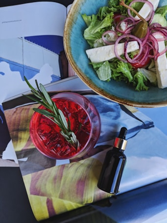 A vibrant green salad served in a blue bowl contains lettuce, sliced onions, feta cheese, and a caper. Next to the bowl, there's a red drink with a sprig of rosemary on top. A small, dark dropper bottle is positioned nearby, all placed on top of an open magazine with colorful pages.