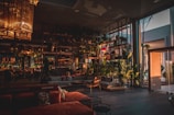 A modern, spacious interior with large glass windows allowing natural light to enter. The room features stylish furniture in warm tones and a variety of indoor plants. Elegant light fixtures hang from the ceiling, adding to the sophisticated atmosphere.