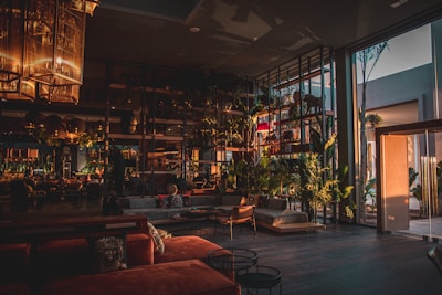 A modern, spacious interior with large glass windows allowing natural light to enter. The room features stylish furniture in warm tones and a variety of indoor plants. Elegant light fixtures hang from the ceiling, adding to the sophisticated atmosphere.