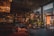 A modern, spacious interior with large glass windows allowing natural light to enter. The room features stylish furniture in warm tones and a variety of indoor plants. Elegant light fixtures hang from the ceiling, adding to the sophisticated atmosphere.