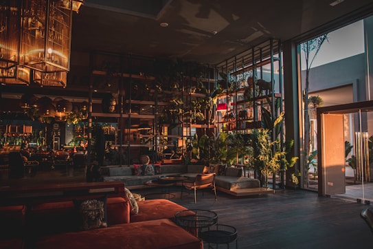 A modern, spacious interior with large glass windows allowing natural light to enter. The room features stylish furniture in warm tones and a variety of indoor plants. Elegant light fixtures hang from the ceiling, adding to the sophisticated atmosphere.