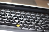 A close-up view of a computer keyboard, featuring a standard layout with English and additional non-Latin characters printed on the keys. The keys are mostly black with white and grey lettering, except for a few keys with orange symbols. The image is slightly out of focus, emphasizing a casual, everyday use context.