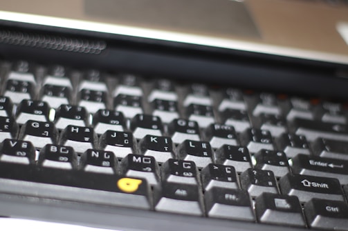 A close-up view of a computer keyboard, featuring a standard layout with English and additional non-Latin characters printed on the keys. The keys are mostly black with white and grey lettering, except for a few keys with orange symbols. The image is slightly out of focus, emphasizing a casual, everyday use context.