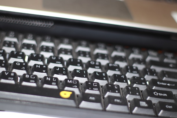 A close-up view of a computer keyboard, featuring a standard layout with English and additional non-Latin characters printed on the keys. The keys are mostly black with white and grey lettering, except for a few keys with orange symbols. The image is slightly out of focus, emphasizing a casual, everyday use context.