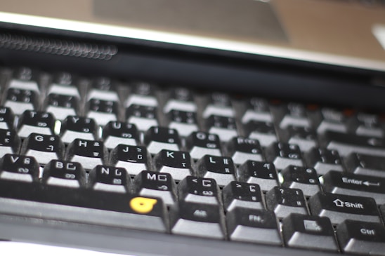A close-up view of a computer keyboard, featuring a standard layout with English and additional non-Latin characters printed on the keys. The keys are mostly black with white and grey lettering, except for a few keys with orange symbols. The image is slightly out of focus, emphasizing a casual, everyday use context.