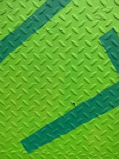 A textured metal surface with a diamond pattern, painted in shades of green. The surface features two diagonal darker green stripes that intersect.