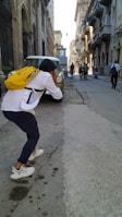 A person with a yellow backpack is taking a photo on a narrow street lined with old buildings. Several people are walking in the distance with some buildings casting shadows on the pavement. The atmosphere suggests an urban setting.