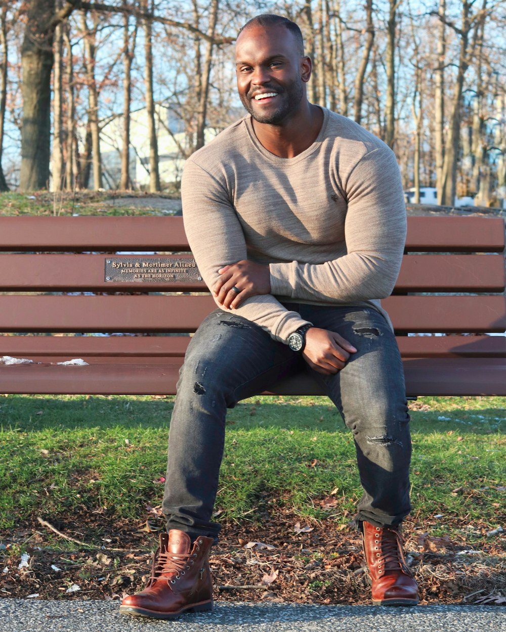 Mann in grauem Pullover und blauer Jeans sitzt auf brauner Holzbank