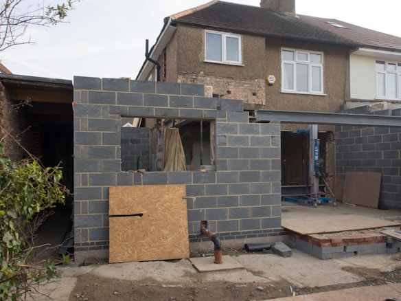 A partially constructed building extends from the back of a house, with dark gray cinder block walls and a window opening. Construction materials, such as boards and metal beams, are visible inside the structure. The original stone siding of the house is visible, and some of the bricks have been exposed. An unfinished pipe is protruding from the ground.