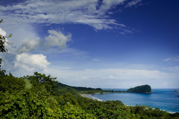 A lush green coastline curves into the distance, bordered by vibrant blue ocean waters. On the horizon, an island rises with dense foliage. The sky is partially cloudy with a clear blue expanse.