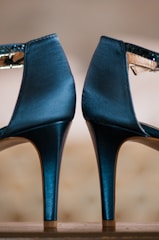 A close-up view of a pair of elegant, shiny high-heeled shoes with ankle straps. The shoes are positioned side by side with their heels and back sections prominently displayed. The background is blurred, which makes the shoes the central focus.