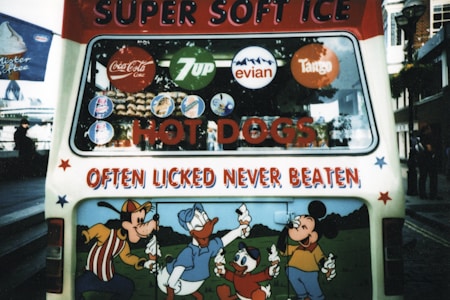 The back of an ice cream van is decorated with colorful cartoon characters and various brand logos. The text 'SUPER SOFT ICE' and 'OFTEN licked NEVER BEATEN' is prominently displayed. Logos for popular beverages such as Coca-Cola, 7Up, Evian, and Tango are visible. The lower panel features cartoon characters enjoying ice cream cones.