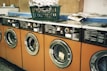 Several orange-fronted washing machines are aligned in a row, each with a circular glass door. On top of one of the machines, there is a green plastic laundry basket filled with clothes. Nearby, a stack of newspapers is placed on a counter.