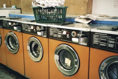 Several orange-fronted washing machines are aligned in a row, each with a circular glass door. On top of one of the machines, there is a green plastic laundry basket filled with clothes. Nearby, a stack of newspapers is placed on a counter.