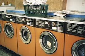 Several orange-fronted washing machines are aligned in a row, each with a circular glass door. On top of one of the machines, there is a green plastic laundry basket filled with clothes. Nearby, a stack of newspapers is placed on a counter.