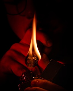 A close-up view of a person lighting a cigar with a metal lighter. The scene is dimly lit with the flame being the primary light source, casting a warm glow on the cigar and the person's fingers holding both the cigar and the lighter.