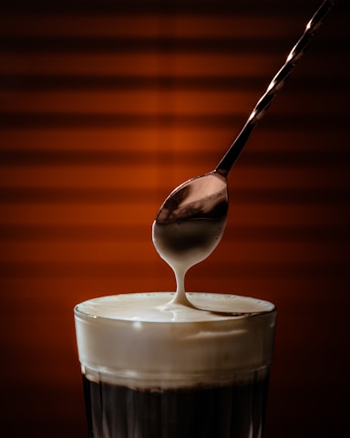 A spoon drizzles creamy liquid into a glass filled with a dark beverage. The background features warm, soft orange tones that create a cozy ambiance.
