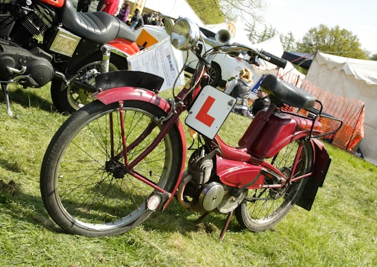 A vintage red moped is parked on grass near a large tent. It has a learner plate attached to the front and is accompanied by a larger motorcycle in the background. People can be seen milling around the area, which appears to be an outdoor event or fair.