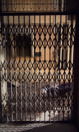 A metal accordion-style gate is closed, casting shadows on the floor. Through the gate, the courtyard of a building is visible, with a parked motorcycle partially obscured. The building walls are textured and aged, and there is debris scattered across the ground.