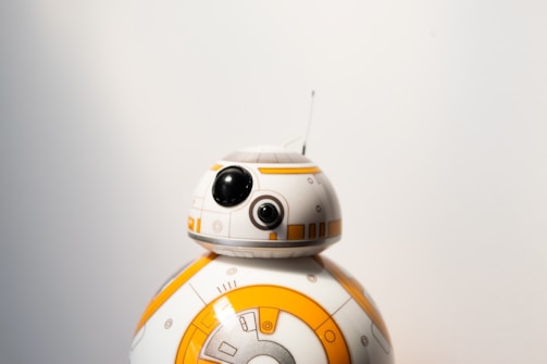A small spherical robot with a round head featuring black and white details and a prominent eye-like lens appears against a plain background. The robot is white with orange circular patterns and markings.