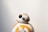 A small spherical robot with a round head featuring black and white details and a prominent eye-like lens appears against a plain background. The robot is white with orange circular patterns and markings.