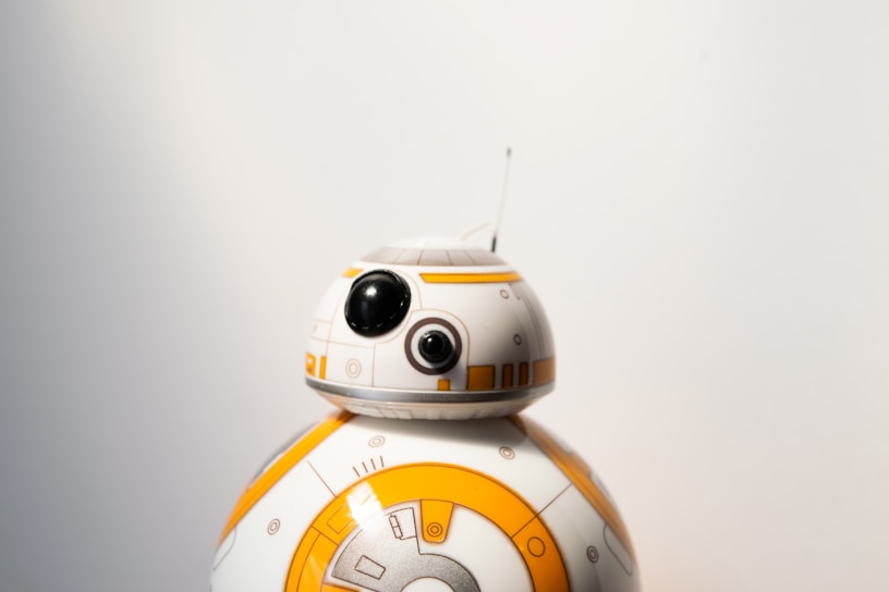 A small spherical robot with a round head featuring black and white details and a prominent eye-like lens appears against a plain background. The robot is white with orange circular patterns and markings.