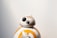 A small spherical robot with a round head featuring black and white details and a prominent eye-like lens appears against a plain background. The robot is white with orange circular patterns and markings.
