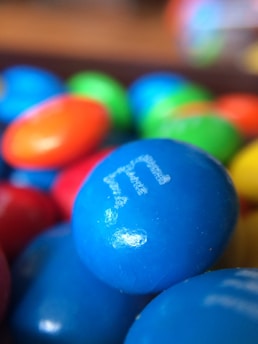 A close-up view of colorful candy-coated chocolate pieces, prominently featuring a blue piece in the foreground with a white letter 'e' printed on it. Surrounding candies include red, orange, and green, with a glossy surface.