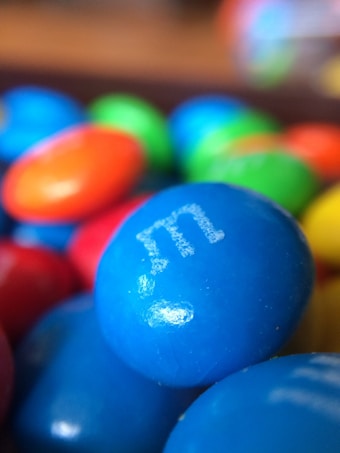 A close-up view of colorful candy-coated chocolate pieces, prominently featuring a blue piece in the foreground with a white letter 'e' printed on it. Surrounding candies include red, orange, and green, with a glossy surface.