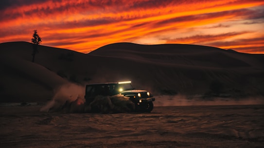 A rugged off-road vehicle traverses a sandy desert landscape as it kicks up a cloud of dust. The dramatic sky is painted with vibrant orange and red hues, suggesting either a sunrise or sunset. The rolling dunes provide a dynamic and adventurous setting.