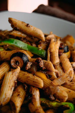 Close-up of a steaming bowl of penne pasta with rich tomato sauce and mixed meats, set against a light beige background with subtle food doodle patterns.
