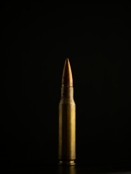 A single brass bullet is displayed vertically against a dark, black background. The lighting highlights its metallic surface, creating a stark contrast between the bullet and the backdrop.