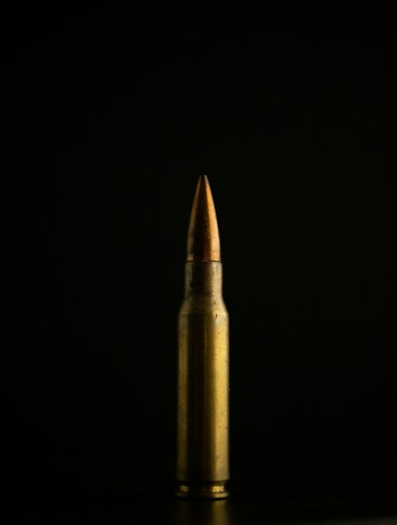 A single brass bullet is displayed vertically against a dark, black background. The lighting highlights its metallic surface, creating a stark contrast between the bullet and the backdrop.