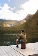 A person sits on a wooden dock by a serene mountain lake, casually fishing. They wear a hoodie, shorts, and a cap, and the rod extends into the water. The backdrop is a picturesque view of tree-covered mountains under a partly cloudy sky.