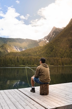A person sits on a wooden dock by a serene mountain lake, casually fishing. They wear a hoodie, shorts, and a cap, and the rod extends into the water. The backdrop is a picturesque view of tree-covered mountains under a partly cloudy sky.