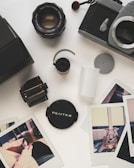 A collection of vintage photography equipment and photos on a light surface. There's a Pentax camera body, a camera lens, film canisters, a lens cap, and several instant photo prints. The photos depict casual scenes, including a person and a dog indoors.