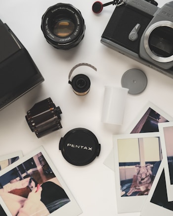 A collection of vintage photography equipment and photos on a light surface. There's a Pentax camera body, a camera lens, film canisters, a lens cap, and several instant photo prints. The photos depict casual scenes, including a person and a dog indoors.