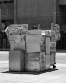 A street food cart with metal siding and several signs advertising items such as hamburgers, hot dogs, and fresh French fries. The cart is situated in an urban environment with a sidewalk and a concrete building in the background. The overall setup is simple and functional, intended for quick service.