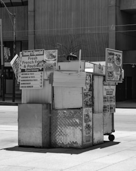 A street food cart with metal siding and several signs advertising items such as hamburgers, hot dogs, and fresh French fries. The cart is situated in an urban environment with a sidewalk and a concrete building in the background. The overall setup is simple and functional, intended for quick service.