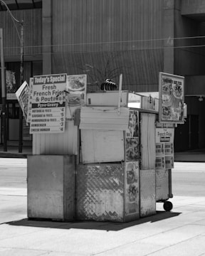 A street food cart with metal siding and several signs advertising items such as hamburgers, hot dogs, and fresh French fries. The cart is situated in an urban environment with a sidewalk and a concrete building in the background. The overall setup is simple and functional, intended for quick service.